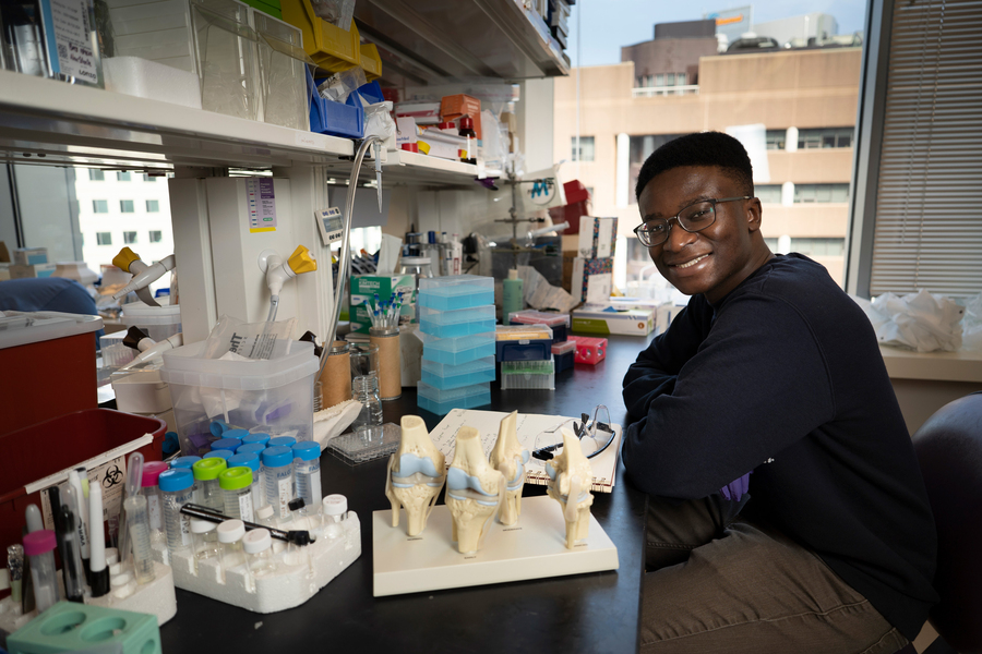Damptey sits at a table in his lab. On the table is a lab notebook, a model of bones, and various plastic boxes and canisters.