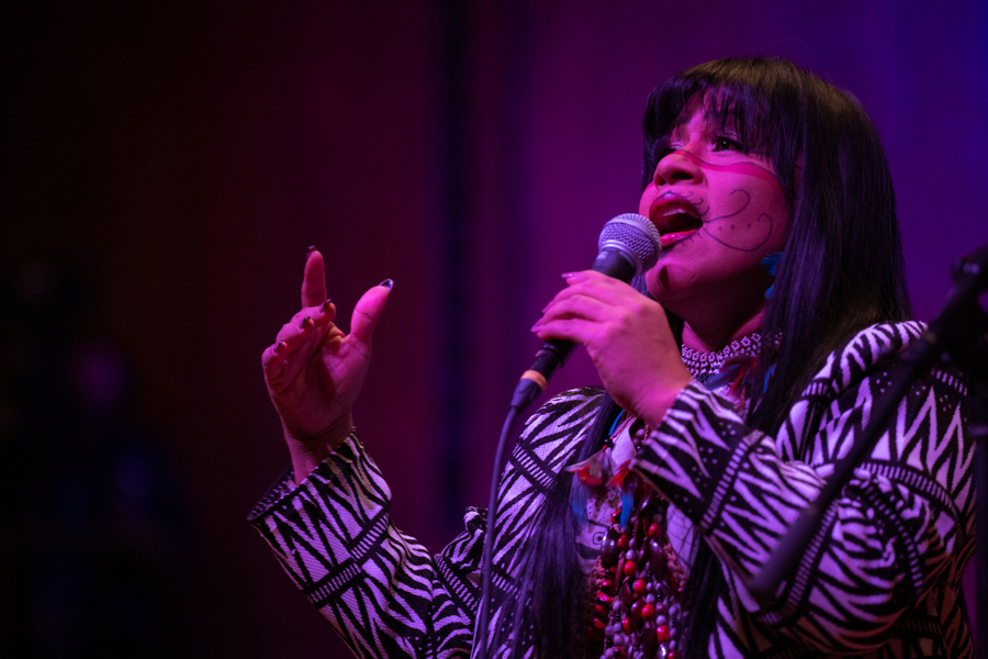 Djuena Tikuna sings while holding a microphone. She is wearing a graphic black-and-white dress and colorful jewelry, and her face is painted with traditional markings.