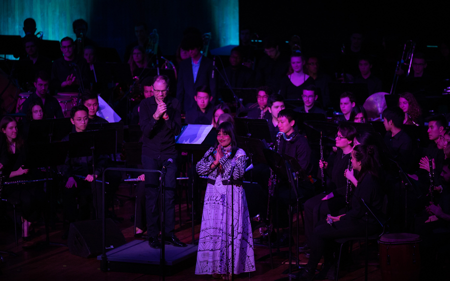 Djuena Tikuna stands on stage in front of many MIT musicians, with her hands pressed together in front of her heart.