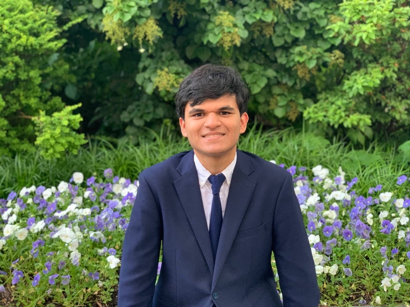 Malhaar Agrawal poses in front of a flower bed, with foliage in the background