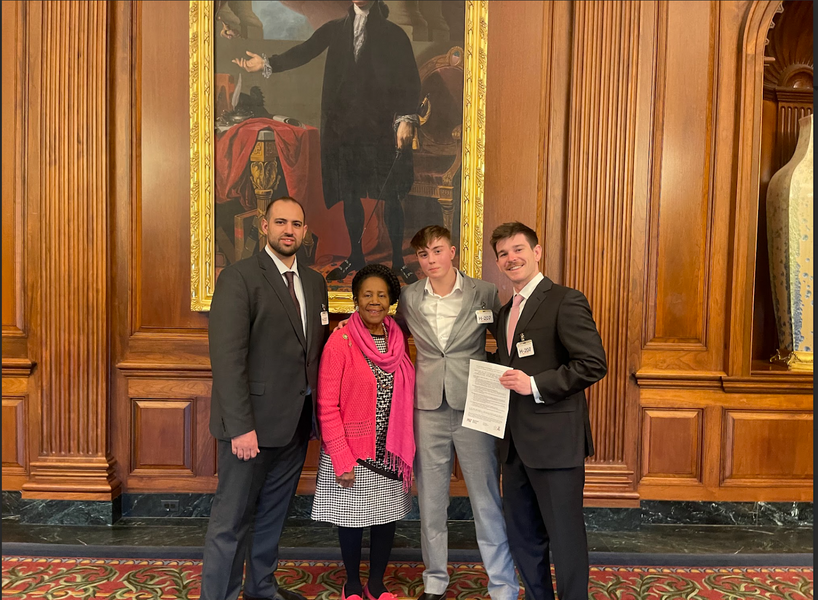 Three MIT students pose with a US representative in front of a wood-paneled wall with columns and a gilt-framed portrait painting of a founder.