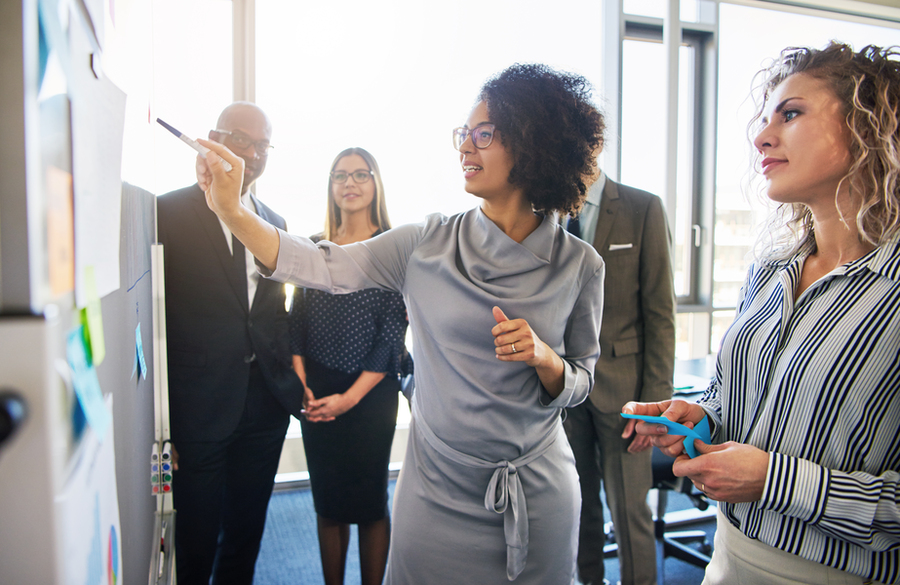 Stock image of a Black woman in a grey dress stands in front of a white board with a pointer while four colleagues look on.