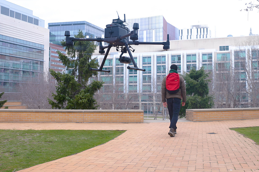 A man seen from behind walks down a brick pathway, followed in the air by a black quadcopter drone