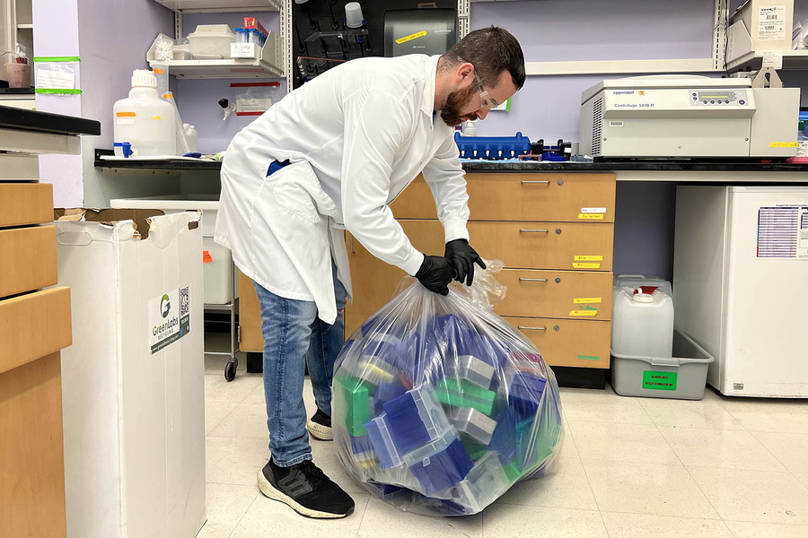 Normand Desrochers in a lab with a white lab coat, black gloves and clear goggles, tying a clear plastic bag of plastics to be recycled.