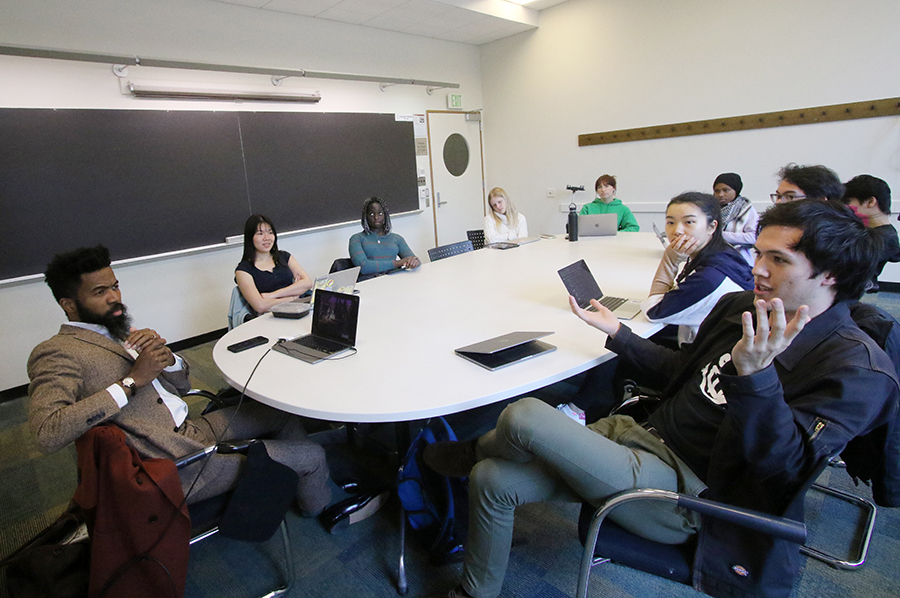 Joshua Bennett speaks to 9 students. They are all seated around an oval table in a small classroom with a blackboard.