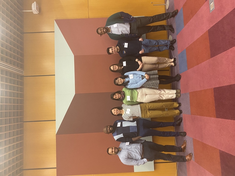 8 people wearing name tags line up for a photo next to a wall featuring a large image of a geometric gemlike shape. The floor carpet is bright stripes of reds and maroon.