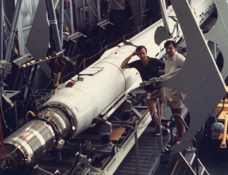 two men next to satellite rocket lying on its side.