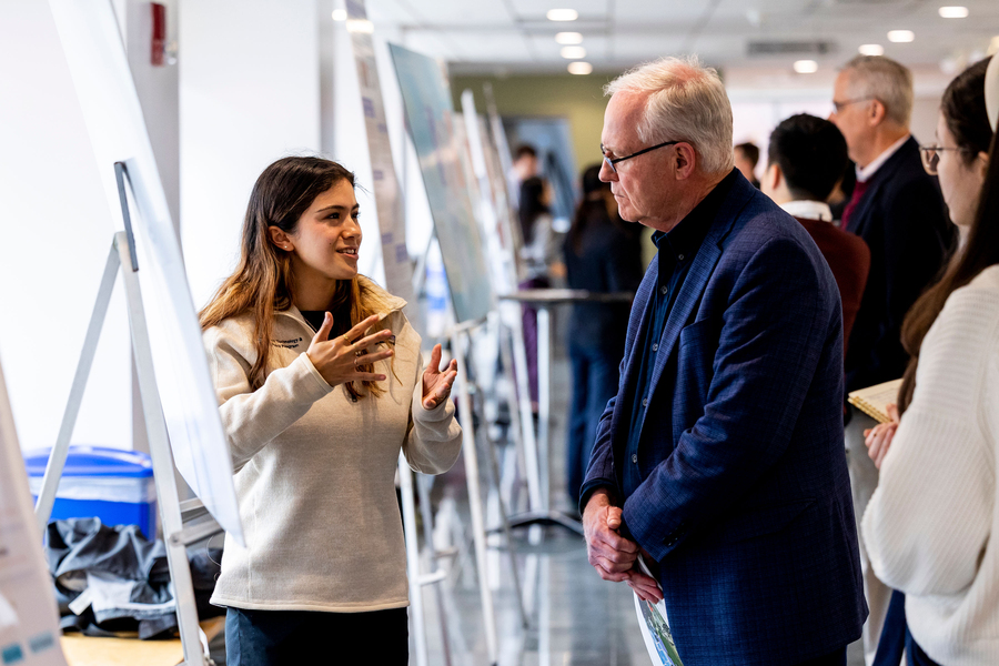 At a poster session with several easels, a smiling MIT student gestures with her hands while a man listens attentively.