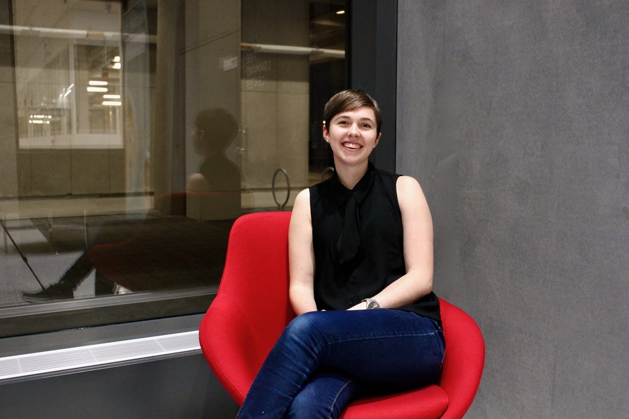 Kelsey Merrill sits in a red chair with a glass window behind her that looks onto some lab space