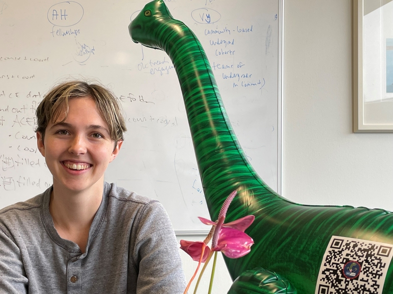 Kelsey Merrill sits in an office with the Vote-a-Saurus inflatable dinosaur in the background.