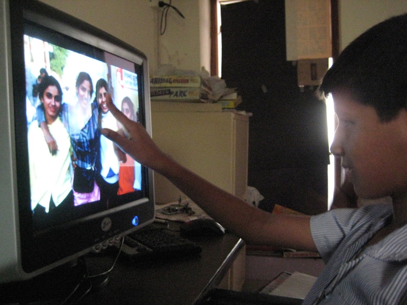 A child is seated in front of a computer screen that has a photo of 4 people smiling. The child’s finger points directly at a person’s face.
