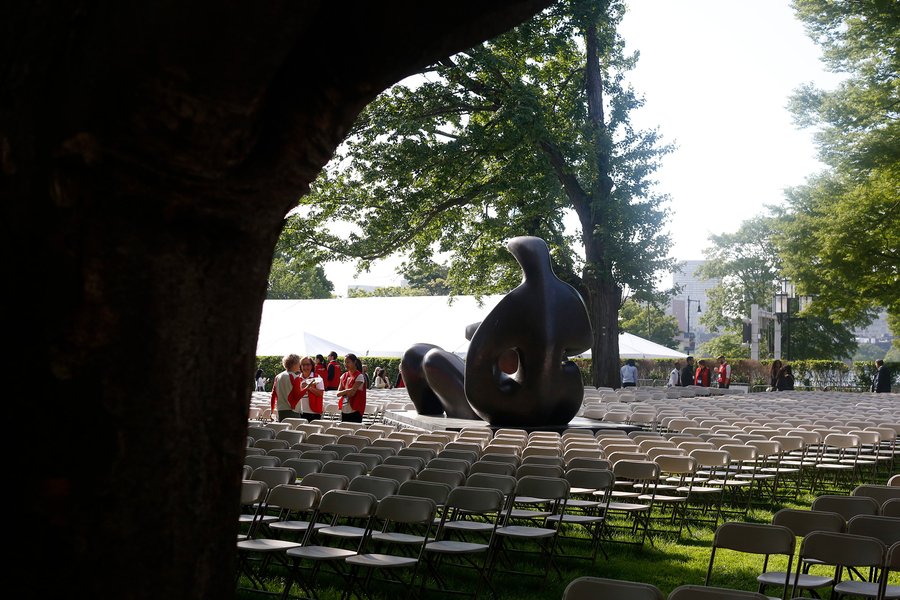 Behind the curvature of a tree trunk in the foreground, hundreds of empty seats in MIT's Killian Court await new graduates and visitors while several volunteers stand amid the seats, next to a large bronze sculpture