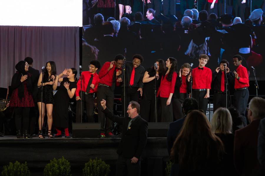 15 singers wear red and black. They are comically reacting to a loud noise made by a singer on the left, laughing while covering their ears.