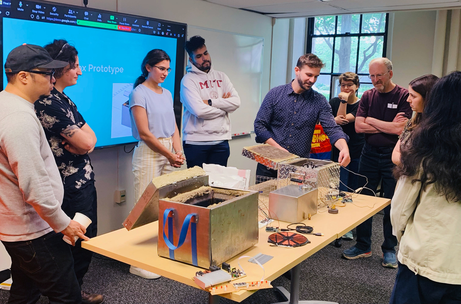9 individuals gather around a table in an MIT classroom to look at two prototype devices, which are small boxes padded with insulation