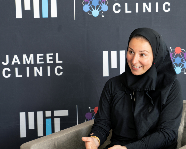 Photo of Marzyeh Ghassemi sitting on a couch and speaking with an MIT Jameel Clinic backdrop behind her.