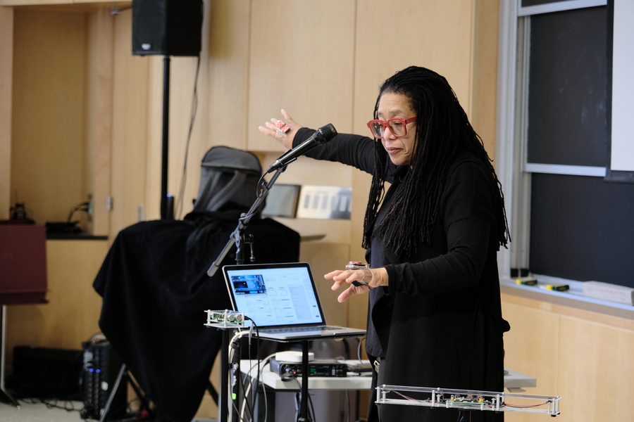 Pamela Z stands at a microphone, gesturing at electronic instruments in a classroom.