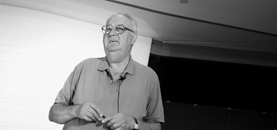 Black-and-white photo of Arnoldo Hax in a polo shirt, standing in front of a blank screen or whiteboard with a microphone clipped to his lapel
