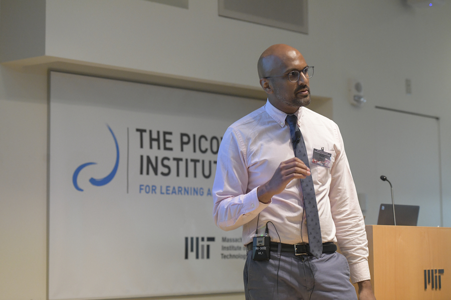 Ravi Raju, in a white shirt, a tie and blue jeans, stands in front of a podium and gestures as he speaks to an audience to the right of the frame.