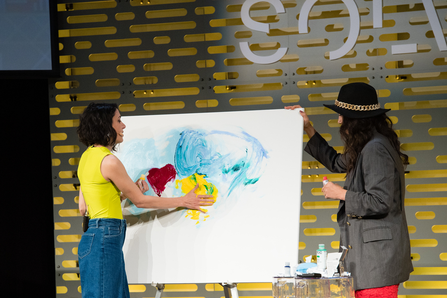 Hala Hanna paints a yellow spot on a large canvas. Aida Murad looks at the painting and braces the canvas from the side. They stand on a stage that is backlit yellow and with the word, "Solve," printed on it.