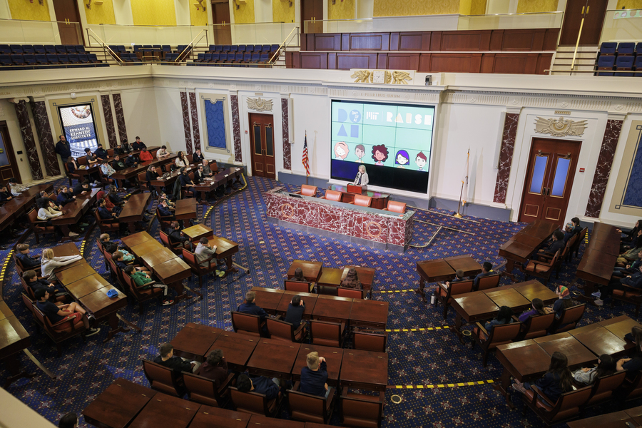 Photo of dozens of middle school girls sitting behind desks in a replica of the U.S. Senate Chambers, listening to MIT President Sally Kornbluth speak at a podium in the center of the room. Behind her is a poster for Day of AI.