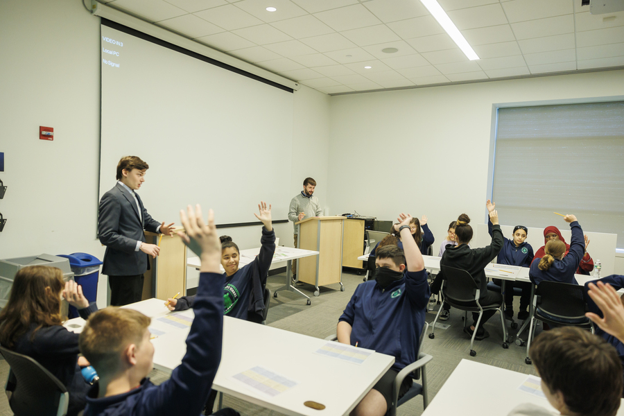Photo of a classroom with two adults in business attire standing at the front. A dozen teenage students sit at several tables with their hands raised.