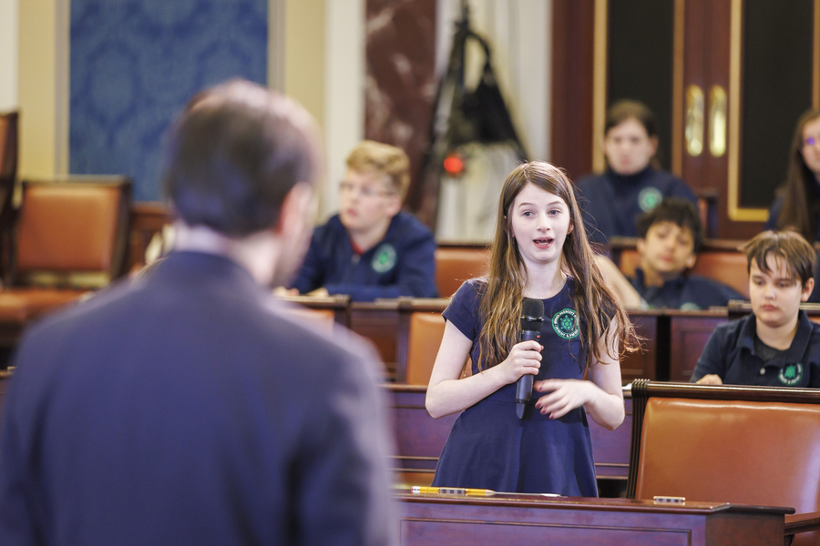 Photo of a middle school girl standing behind a desk in a replica of the U.S. Senate Chambers, speaking into a microphone. Several students and adults look on.