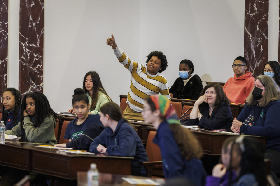 Photo of a middle school boy standing behind a desk next to half-a-dozen seated classmates in a replica of the U.S. Senate Chambers. His thumb is pointing up for a "Yay" vote.