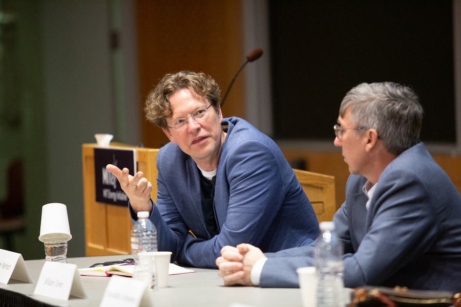 Christoph Reinhart speaks while sitting on a panel. Another panelist beside him looks on. There is a lecturn and a blackboard in the background.