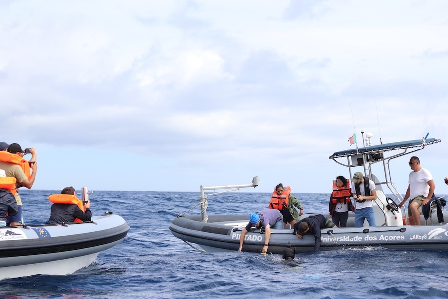 Students on two small boats wear orange life-vests. On one of the boats, two of the students are reaching over into the water. 