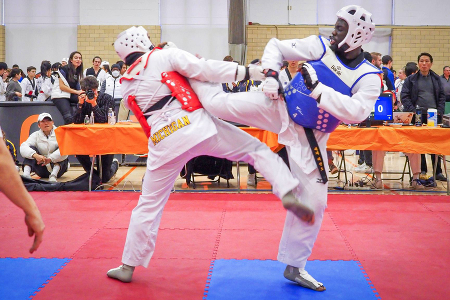 At a competition, Moctar, on right and in blue, kicks dramatically at a competitor in red. The competitor’s uniform says “Michigan.”