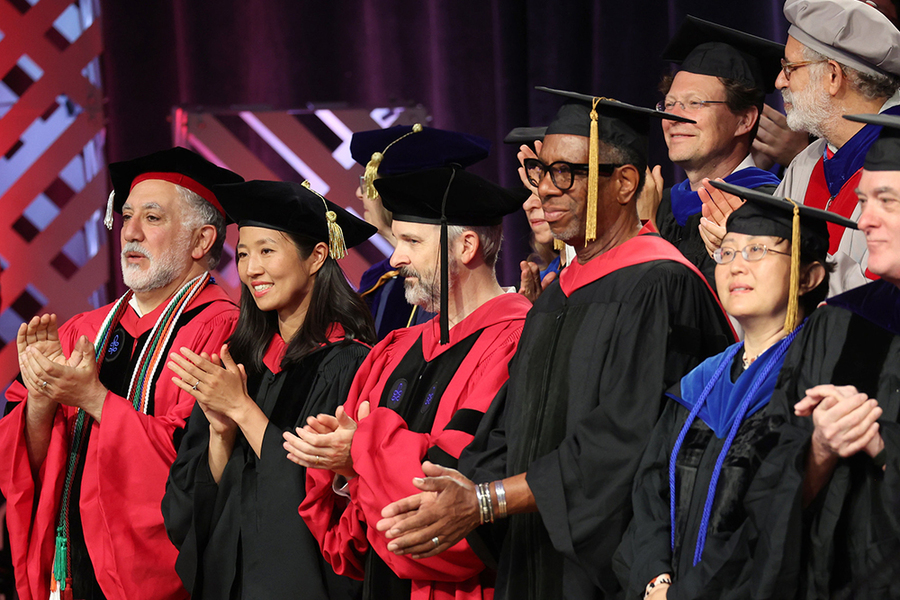 Eight men and women in academic regalia applauding on a stage indoors