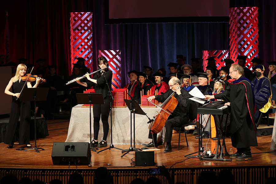 A quartet plays music on a wooden floor while audience members, seated in academic regalia, look on from behind