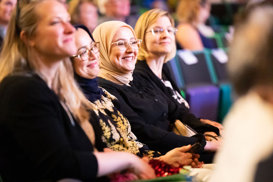 Closeup of four women, two wearing headscarves, smiling warmly while seated in an auditorium