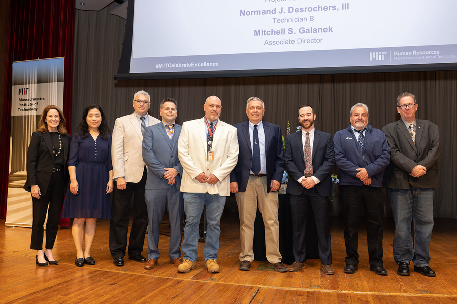 Nine adults stand in a row on a stage with a banner reading “Massachusetts Institute of Technology” behind them.