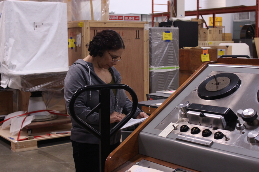Gloria Martinez reviews a to-do list. She stands to the left of a large 20th century machine that is nearly as tall as she is. Behind her are large crates and boxes packed in plastic with labels on them.