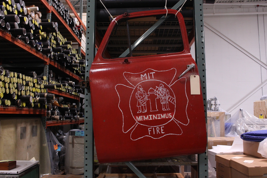 Red door of a firetruck is suspended with ropes in a museum collections warehouse. On the door is a hand-painted insignia mimicking MIT's logo with two firefighters facing away from each other and a hydrant between them along with the words “MIT MEMINIMUS FIRE." Metal shelves with hundreds of long cylinders are at left. At right are a bunch of boxes and other items covered with plastic.