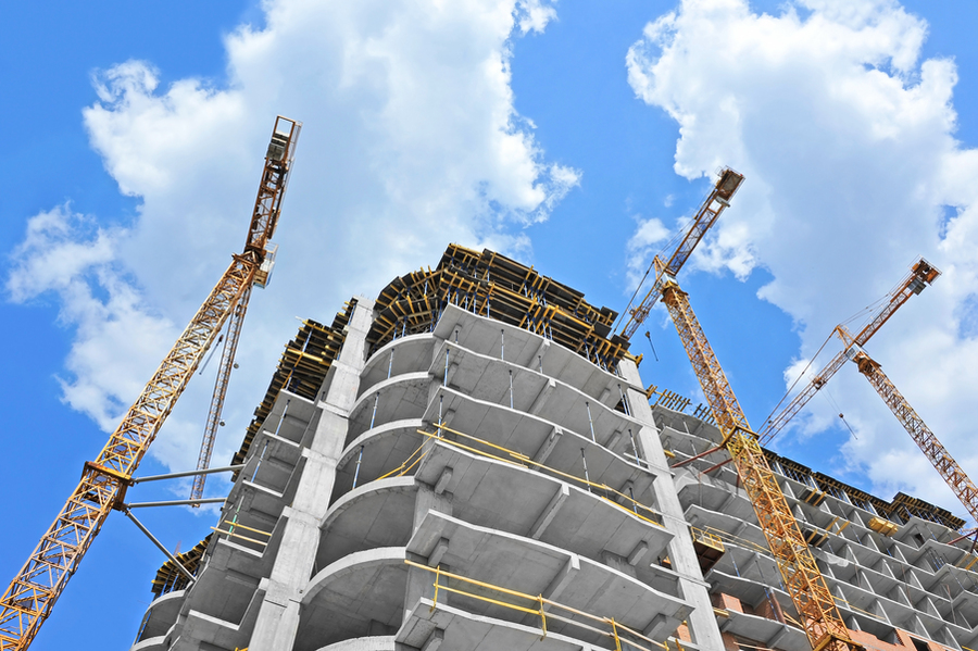 Photo of a tall concrete slab building under construction. The view is looking up against a blue sky with puffy white clouds.