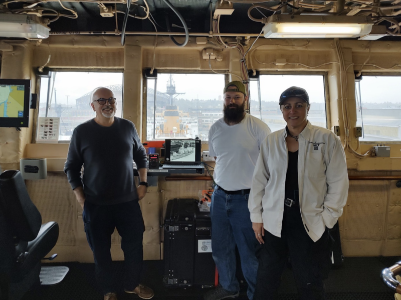Paul Metzger, Michael Emily, and Amna Greaves stand inside a ship's superstructure, with a computer monitor showing imagery behind them. The ocean is also seen behind them through windows.