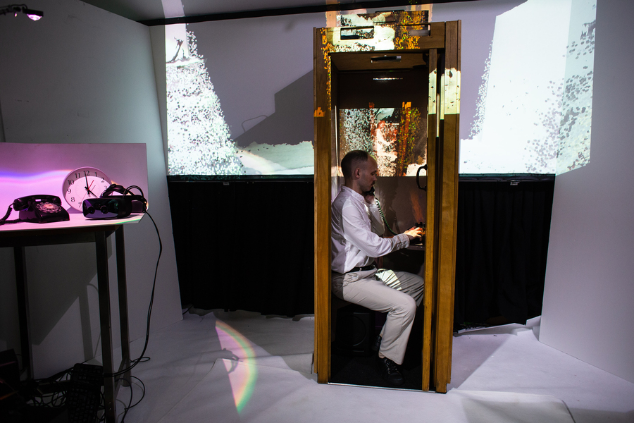 A man sits in a wooden phone booth in front of a multimedia light design