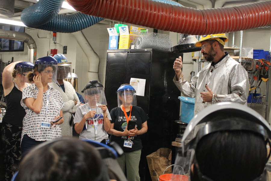 A man in a hard hat and mylar jacket lectures five teachers, all wearing hard hats with face shields
