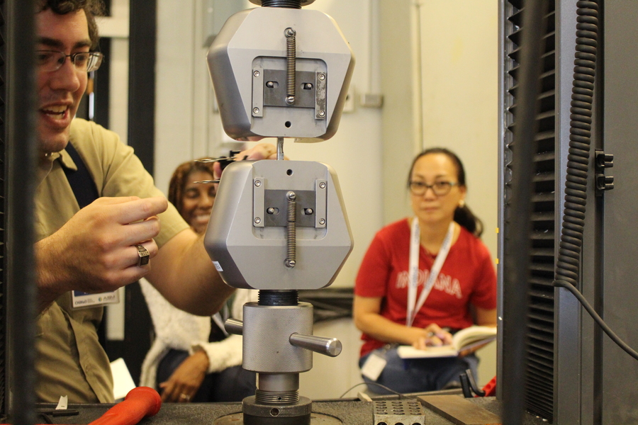 Shaymus Hudson demonstrates a piece of lab equipment to two soft-focus women seated in the background