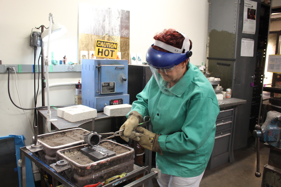 Brenna Toblan, wearing heavy protective gear, pours liquid metal into a mold in a lab space