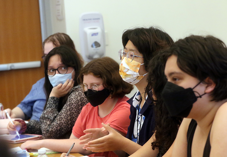 Four college students wearing masks sit at a table. One is gesturing with her hands.