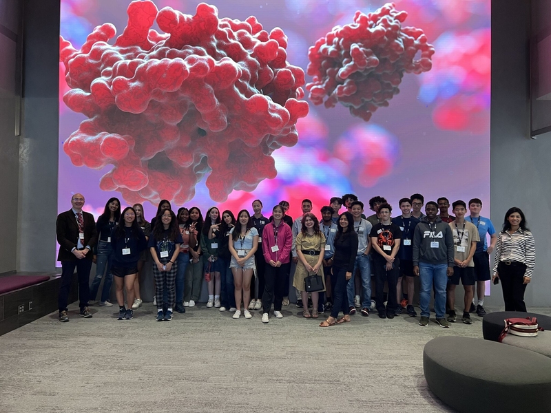 Photo of about 30 high school students posing in front of a large screen filled with illustrations of red molecules.