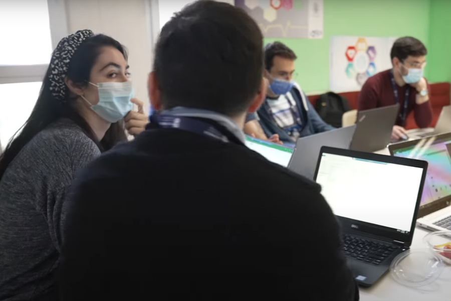 Photo of the back of someone's head, sitting at a table in a classroom with several out-of-focus individuals wearing face masks and using laptops