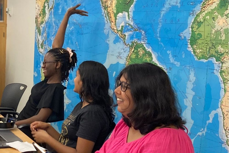 Three women sit at a table with a world map on the wall behind them. One has her hand raised.