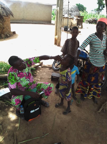 A man in a brightly colored shirt and pants squats behind a black bag. He is tipping a dose of vitamin A into the mouth of a child who is standing with their head tilted upward.
