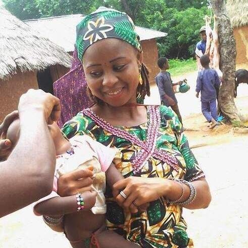 A woman in a bright green dress and headwrap holds a young child whose head is tilted back. The hand of someone outside the frame obscures the child’s face.