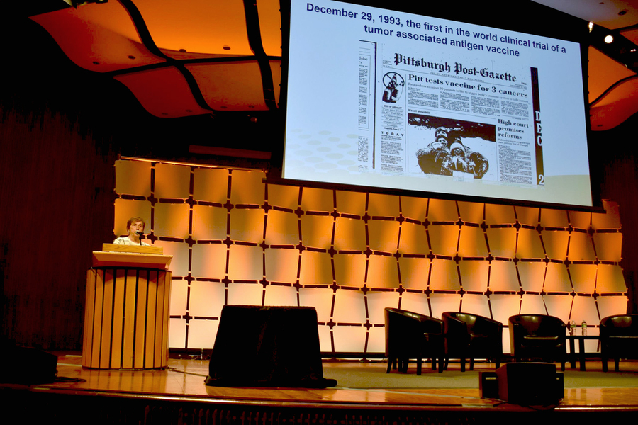 A woman speaks at a lectern on a stage, displaying behind her a slide of a newspaper with the headline "Pitt tests vaccine for 3 cancers"