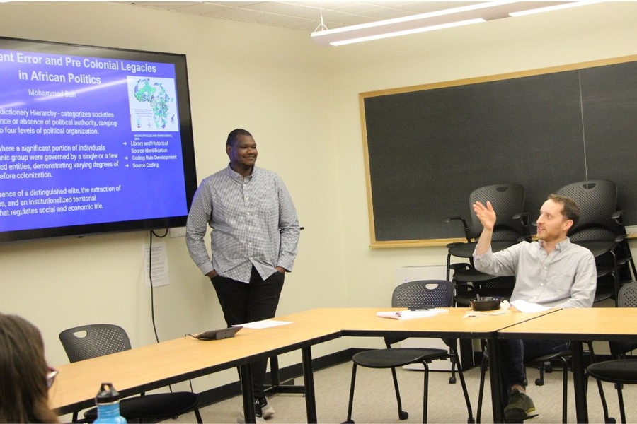A man stands in front of a screen with his hands in his pockets, as a seated man gestures while speaking                  